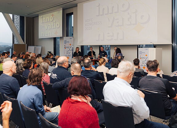 Blick über die Köpfe zahlreicher sitzender Zuschauer im Saal auf eine Bühne. Dort diskutieren vier Personen bei einer Podiumsdiskussion. Im Hintergrund zeigen Leinwände den Schriftzug ADFC Kongress 2026 Innovationen.