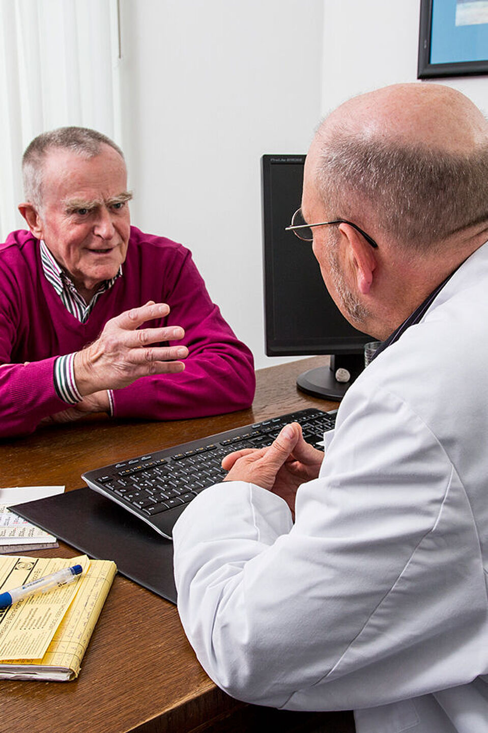 Patientengespräch Ein Arzt am Schreibtisch ist von schräg hinten zu sehen, wie er mit einem Patienten spricht.