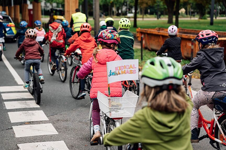 Kidical Mass Mehrere Kinder und Erwachsene fahren bei einer Kidical Mass gemeinsam mit Fahrrädern und Helmen auf einer Straße. Ein Kind trägt eine Weste und hat ein Schild mit „Kidical Mass“ am Fahrradkorb.