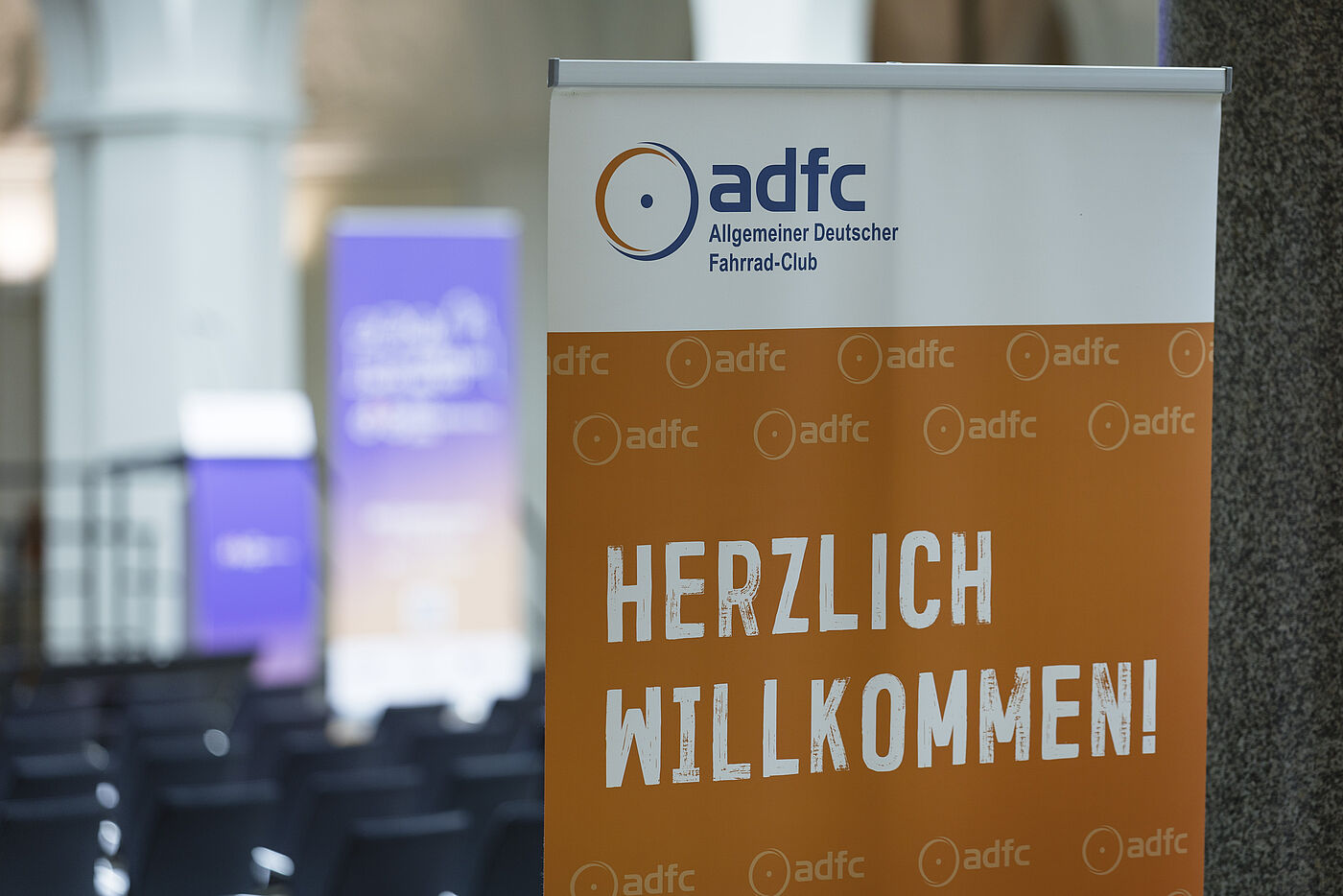 ADFC-Willkommensschild Oranges ADFC-Banner mit weißer Schrift "HERZLICH WILLKOMMEN!" und ADFC-Logo, im Hintergrund unscharf lila Banner und Veranstaltungsbereich